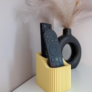 May include: A yellow, ribbed remote control tidy holding two black remotes. The holder has a rectangular shape with vertical grooves. A black vase and decorative feathers are in the background.