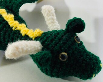May include: Green crocheted dragon with yellow and white accents. The dragon has large black eyes and a small yellow nose.