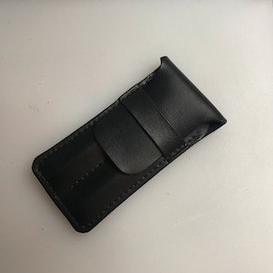 May include: A black leather pen case with a flap closure. The case is rectangular with a stitched border and a loop detail. The leather has a smooth, polished finish. The case is designed to hold a single pen or pencil.