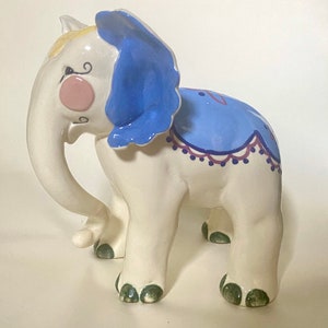 May include: A white ceramic elephant figurine with blue and purple painted details. The elephant has a blue and white patterned blanket on its back and green painted feet.