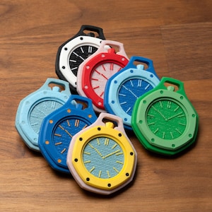 Op de afbeelding: Een verzameling kleurrijke, gestileerde objecten in de vorm van horloges. Elk horloge heeft een ander kleurenschema, waaronder zwart, rood, blauw, groen en geel. De wijzerplaten van de horloges hebben een textuur met eenvoudige uurmarkeringen en wijzers. De objecten zijn gerangschikt op een houten oppervlak.