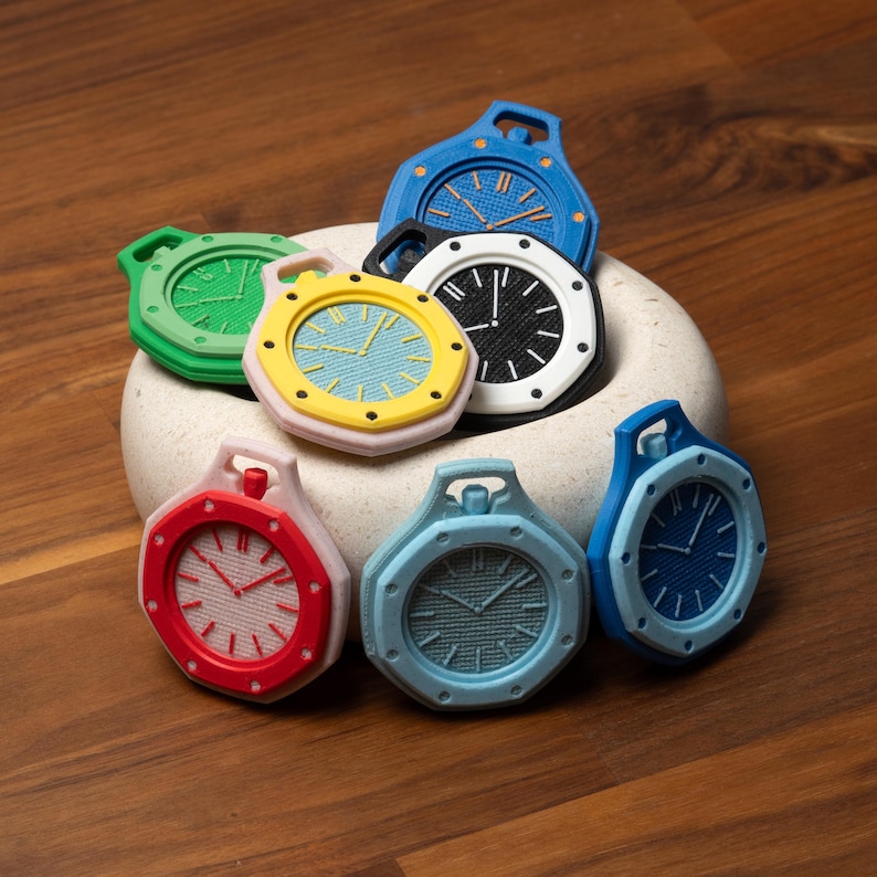 May include: A collection of colorful, miniature clock-shaped objects. The clocks feature various colors, including red, green, blue, and yellow, with contrasting clock faces. The objects are arranged in a white, rounded container, set against a wooden surface.