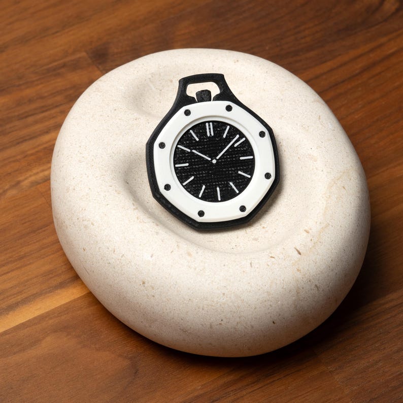 May include: A black and white watch with a textured black face and white accents. The watch is set on a light-colored, rounded stone, with a dark wooden background.