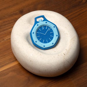 May include: A blue and light blue pocket watch design. The watch has a round face with Roman numerals and hands indicating the time. The watch is set on a white, oval-shaped stone.