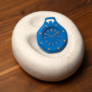 May include: A blue pocket watch with orange accents rests on a white, textured stone. The watch features a detailed face with hands and markers. The watch is a modern take on a classic timepiece.