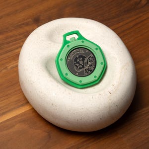 May include: A green, octagonal-shaped pendant with a detailed, black inner design, resting on a white, rounded stone. The pendant has a loop at the top. The stone has a circular indentation. The design inside the pendant is intricate.