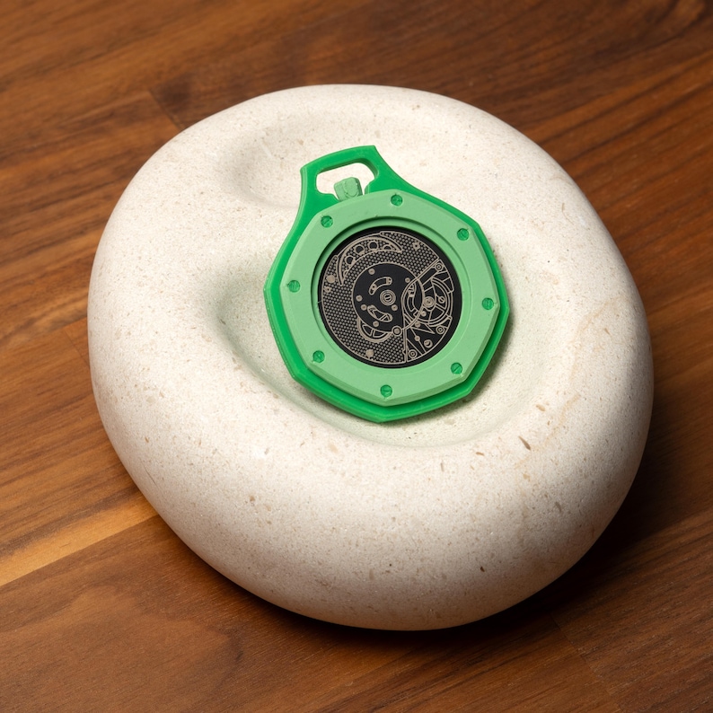 May include: A green, octagonal-shaped pendant with a detailed, black inner design, resting on a white, rounded stone. The pendant has a loop at the top. The stone has a circular indentation. The design inside the pendant is intricate.