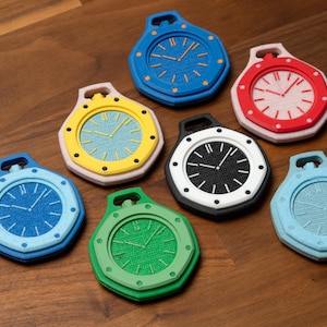 May include: A collection of seven colorful, stylized clock-shaped objects. Each clock features a different color scheme, including blue, red, yellow, green, black, and light blue. The clocks have a textured face and a loop at the top.