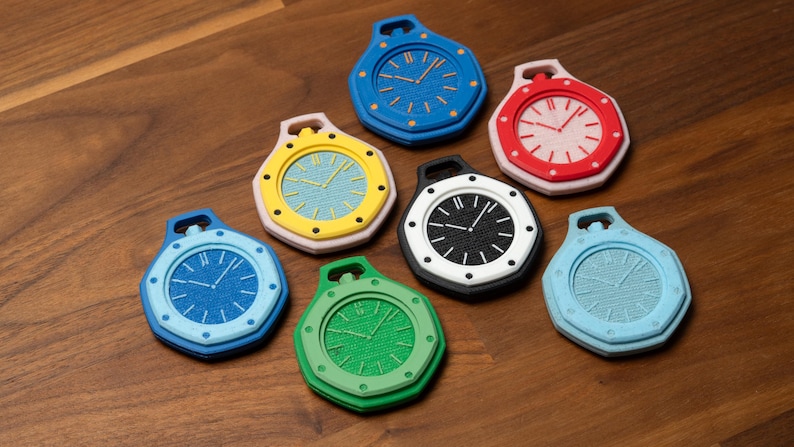 May include: A collection of seven colorful, stylized clock-shaped objects. Each clock features a different color scheme, including blue, red, yellow, green, black, and light blue. The clocks have a textured face and a loop at the top.