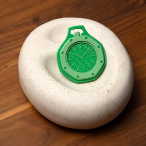 May include: A green watch pendant with a circular face and Roman numeral hour markers. The pendant is set on a white, textured, oval-shaped stone, against a dark wood surface. The watch has a detailed face and a loop at the top.