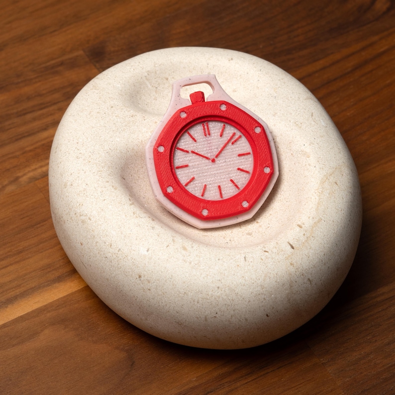 May include: A red and white decorative clock pendant with a detailed face, resting on a light-colored, textured stone. The clock has a red bezel and a white face with black hands and markers. The stone sits on a wooden surface.