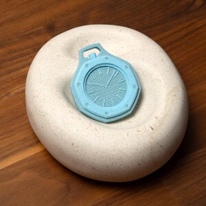 May include: A light blue, octagonal-shaped pendant resembling a pocket watch, resting on a smooth, off-white stone. The pendant features a detailed clock face design and a small loop at the top. The stone sits on a wooden surface.