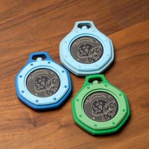 May include: Three colorful, hexagonal-shaped coasters with a circular, detailed mechanical design in the center. The coasters are in shades of blue and green, with a dark background. The coasters have a loop at the top.