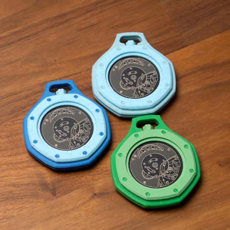 May include: Three colorful, hexagonal-shaped coasters with a circular, detailed mechanical design in the center. The coasters are in shades of blue and green, with a dark background. The coasters have a loop at the top.