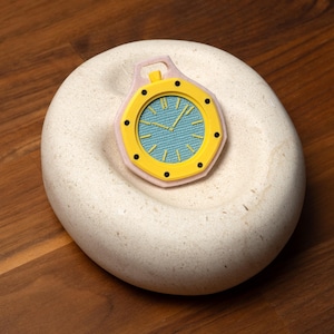 May include: A yellow and black watch with a light blue face, set on a white, rounded stone. The watch has a pink border and is placed on a wooden surface. The watch hands point to the time.