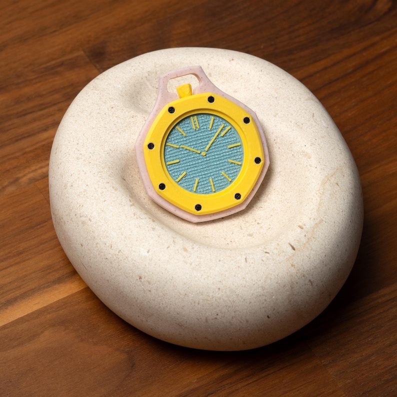 May include: A yellow and black watch with a light blue face, set on a white, rounded stone. The watch has a pink border and is placed on a wooden surface. The watch hands point to the time.