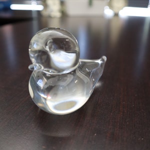Peut inclure: Une figurine en verre transparent en forme de petit canard. L'objet présente un corps lisse et arrondi, une tête définie avec un petit bec et une queue pointue. Le verre est transparent et reflète la lumière, créant des reflets sur sa surface.