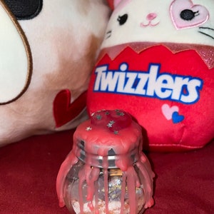 May include: A small glass jar with a silver lid, covered in pink wax that is dripping down the sides. The jar is filled with small, colorful objects. A red plush toy with the word "Twizzlers" is in the background.