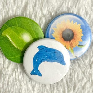 May include: Three round buttons with nature-themed designs. One button features a green leaf, another a blue dolphin, and the third a sunflower against a blue sky. The buttons are on a white, textured surface.