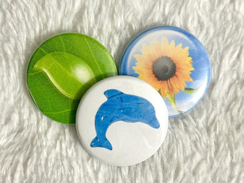 May include: Three round buttons with nature-themed designs. One button features a green leaf, another a blue dolphin, and the third a sunflower against a blue sky. The buttons are on a white, textured surface.