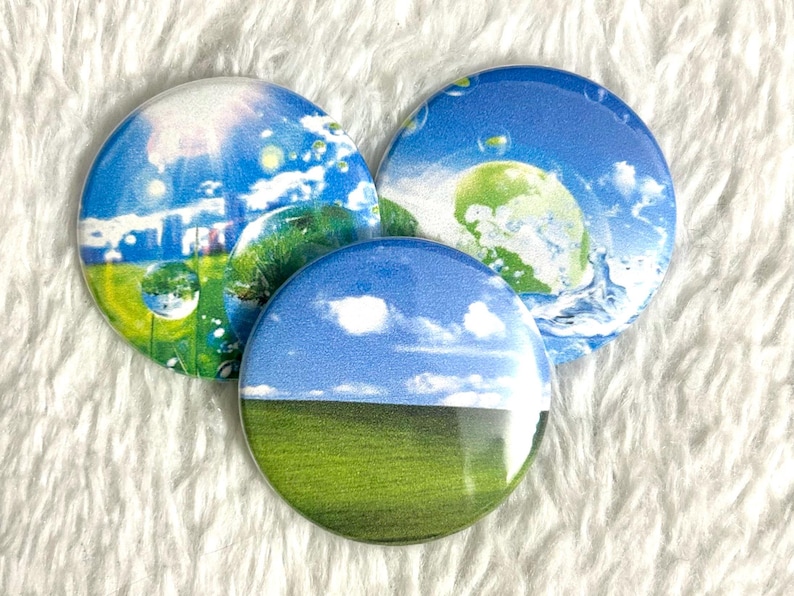 May include: Three round buttons with landscape designs. One button features a blue sky with white clouds over a green field. The other two buttons have similar designs with blue skies, green fields, and water droplets.