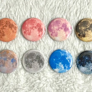 May include: Eight circular, glossy buttons featuring moon phase designs. The buttons display a range of colours, including orange, pink, purple, brown, grey, blue, and black. The buttons are arranged in two rows of four.
