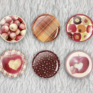 May include: A collection of six round decorative magnets. The designs include apples, plaid patterns, and a star pattern. One magnet features a heart-shaped cutout in an apple. The color palette includes red, brown, and cream.