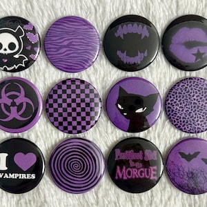 May include: A collection of twelve round black and purple buttons with various designs. The designs include a skull, zebra stripes, vampire fangs, lips, a biohazard symbol, a checkerboard, a cat, leopard print, the text "I ❤️ VAMPIRES", a spiral, the text "Prettiest Girl in the MORGUE", and bats.