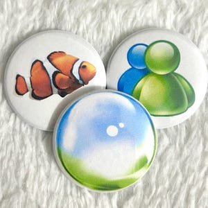 May include: Three round white buttons with colorful designs. One button features a clownfish, another has two figures in blue and green, and the third has a blue and green bubble design. The buttons are on a white, textured surface.
