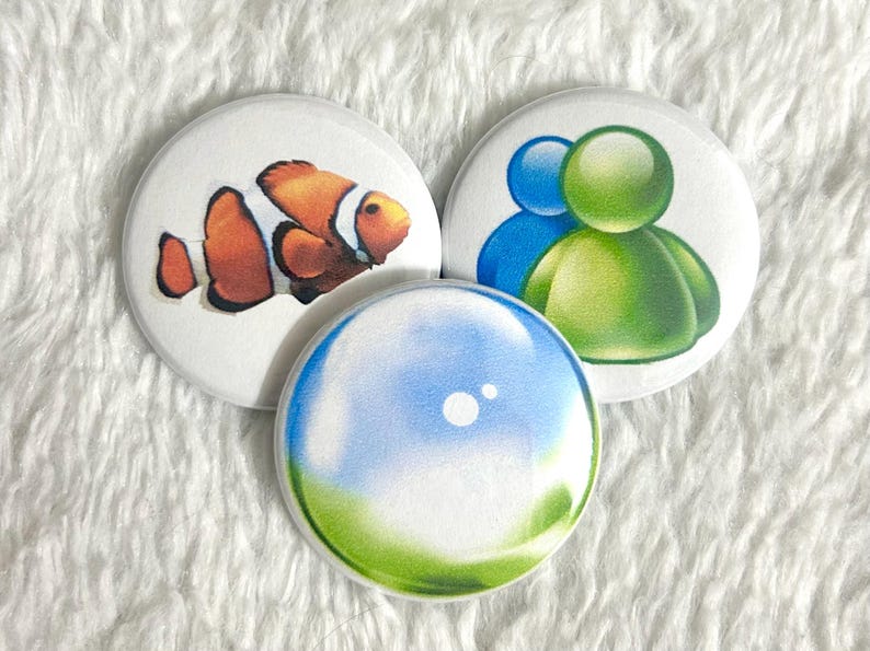 May include: Three round white buttons with colorful designs. One button features a clownfish, another has two figures in blue and green, and the third has a blue and green bubble design. The buttons are on a white, textured surface.
