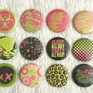 May include: A collection of twelve round pinback buttons with various designs. The buttons feature neon colors and graphics, including a biohazard symbol, abstract patterns, cartoon characters, and the text "SLEEP ALL DAY PARTY ALL NIGHT."
