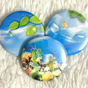 May include: Three round, blue decorative magnets with aquatic designs. One features green leaves, another a goldfish, and the third a colorful coral reef with fish. The magnets are arranged on a white, textured surface.