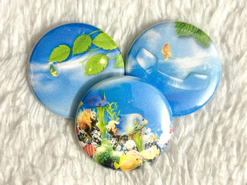 May include: Three round, blue decorative magnets with aquatic designs. One features green leaves, another a goldfish, and the third a colorful coral reef with fish. The magnets are arranged on a white, textured surface.