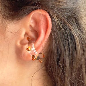 以下が含まれることがあります： ゴールドカラーのハート型アクセントが付いたクリアイヤーカフ。 イヤーカフは耳に巻き付き、ユニークでモダンな外観を作り出します。 このジュエリーは、茶色の髪の人の耳に着用されています。