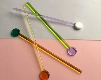 May include: Four glass stirring sticks with colored glass ends. The sticks are green, purple, orange, and green. The ends are round and the sticks are lying on a pink and white surface.