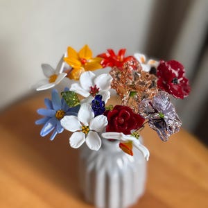 May include: A small, white, ribbed vase holds a colorful bouquet of glass flowers. The flowers include white, yellow, red, blue, and purple blooms. The vase sits on a wooden surface.