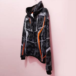 Pode incluir: Um casaco com capuz e z&iacute;per completo com um padr&atilde;o de camuflagem em tons de cinza e preto, acentuado com desenhos geom&eacute;tricos laranja e branco. O casaco tem capuz e punhos pretos e est&aacute; pendurado em um cabide de metal.