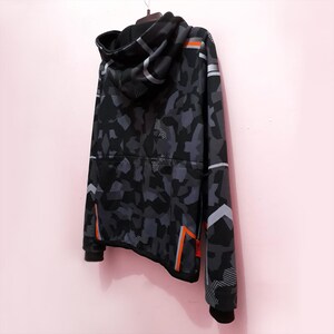 Pode incluir: Um moletom com capuz camuflado preto e cinza com capuz com cord&atilde;o. O moletom tem listras de destaque laranja e branco nas mangas e laterais. A roupa est&aacute; pendurada em um cabide em um fundo rosa.