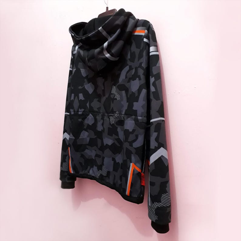 Pode incluir: Um moletom com capuz camuflado preto e cinza com capuz com cord&atilde;o. O moletom tem listras de destaque laranja e branco nas mangas e laterais. A roupa est&aacute; pendurada em um cabide em um fundo rosa.