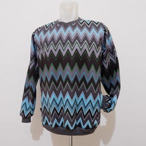 May include: A long-sleeved sweatshirt featuring a repeating chevron pattern in shades of light blue, dark grey, black, and green. The garment has a crew neckline and ribbed cuffs and hem in a solid dark grey colour, displayed on a mannequin.