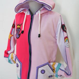 Puede incluir: Sudadera con capucha rosa y magenta con cremallera, capucha y orejas de oso. La chaqueta presenta un diseño colorido con una corona y el número 12 en la parte delantera. Las mangas tienen gráficos de arcoíris y nubes. La sudadera tiene cremallera completa y bolsillos.