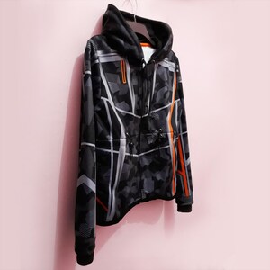 Pode incluir: Um casaco com capuz com z&iacute;per com padr&atilde;o de camuflagem preto e cinza com detalhes em branco e laranja. O casaco tem um cord&atilde;o na cintura e um capuz preto. O casaco est&aacute; pendurado num cabide sobre um fundo rosa.