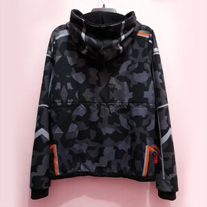 Pode incluir: Casaco com capuz camuflado preto e cinza com gola alta. O casaco apresenta faixas de destaque laranja e branco e uma bainha inferior preta. O padr&atilde;o &eacute; uma mistura de formas geom&eacute;tricas.