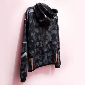 Pode incluir: Um moletom com capuz camuflado preto e cinza com z&iacute;per completo. O design inclui formas geom&eacute;tricas e detalhes em laranja nas mangas e laterais. O moletom est&aacute; pendurado em um cabide.