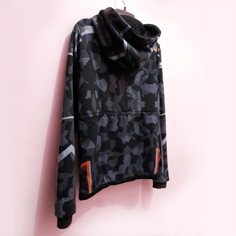Pode incluir: Um moletom com capuz camuflado preto e cinza com z&iacute;per completo. O design inclui formas geom&eacute;tricas e detalhes em laranja nas mangas e laterais. O moletom est&aacute; pendurado em um cabide.