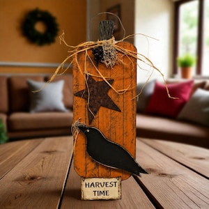 May include: A vertical orange wooden pumpkin-shaped sign with a distressed finish, featuring a dark brown metal star, a black bird silhouette, and a small rectangular base plate with the printed text HARVEST TIME, accented with raffia and berries.