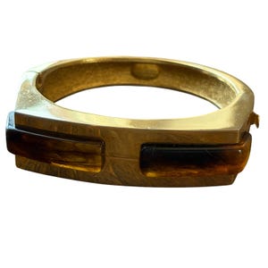 Puede incluir: Una pulsera dorada con bisagras y detalles rectangulares color ámbar. La pulsera tiene forma cuadrada y cierre de broche. Los detalles ámbar son una mezcla de tonos marrones y negros.