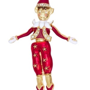 Könnte beinhalten: Eine Brosche aus goldfarbenem Metall in Form eines Elfen, mit roter Emaille-Kleidung mit goldenen Sternakzenten, einem weißen Kragen und weißen Handschuhen. Der Elf trägt eine rote Mütze und eine mit klaren Strasssteinen verzierte Weste sowie ein goldenes Gesicht.