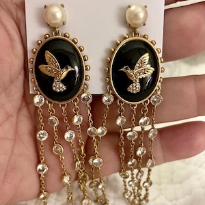 Puede incluir: Pendientes dorados con un centro ovalado negro con un diseño de colibrí dorado y detalles de cristal transparente. Los pendientes tienen un perno de perla y cadenas colgantes con cristales transparentes. La marca "kate spade NEW YORK" es visible.