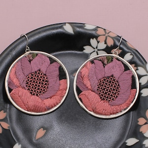 May include: A pair of round earrings featuring embroidered floral designs in shades of pink and purple. The earrings have silver-colored metal frames and hooks. The earrings are displayed on a black plate with a floral pattern.